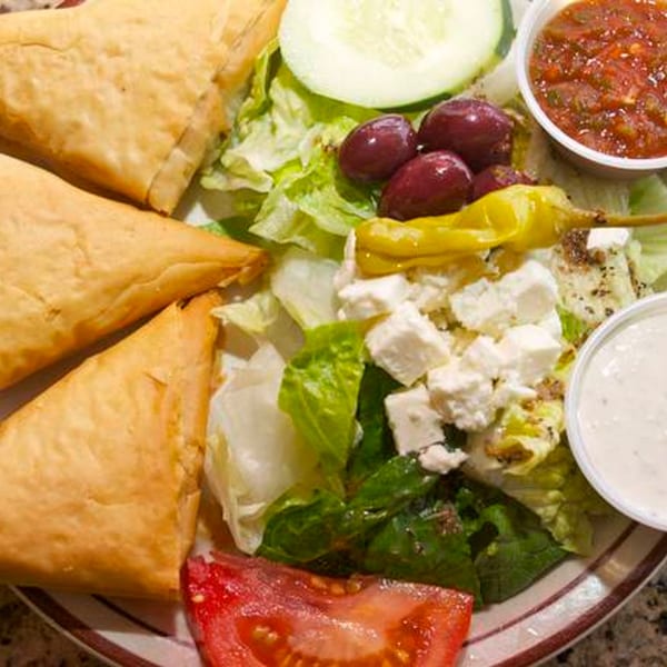 Three golden pastries, a fresh Mediterranean salad with feta, olives, and dressings on a plate.