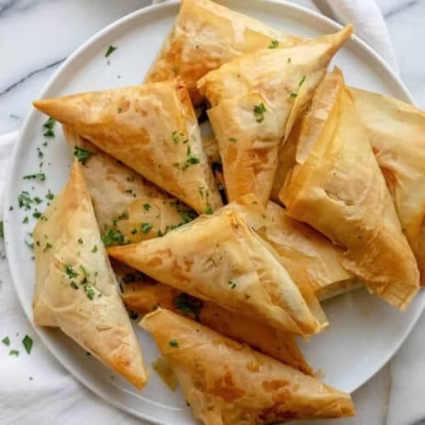 A white plate piled high with golden-brown, triangular spanakopita pastries, garnished with fresh parsley.