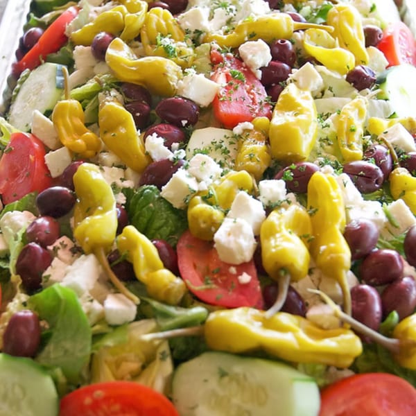 A vibrant Greek salad with lettuce, tomatoes, cucumbers, feta, olives, and yellow pepperoncini.