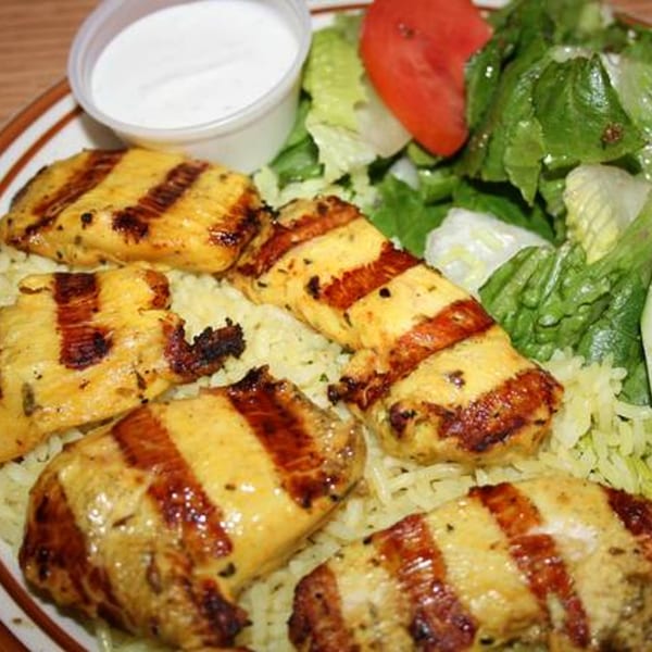 Grilled chicken pieces with rice, a side salad, and a cup of white sauce on a plate.