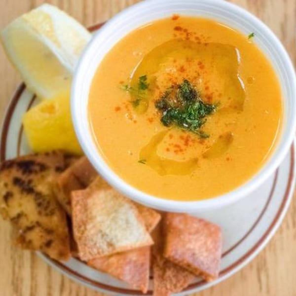 A bowl of orange Mediterranean lentil soup with parsley, olive oil, lemon, and pita bread.
